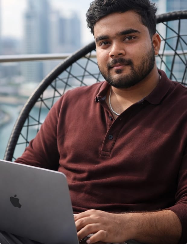 Aditya seated in a woven chair on a high-rise balcony, working on a laptop, with a waterfront skyline behind him.